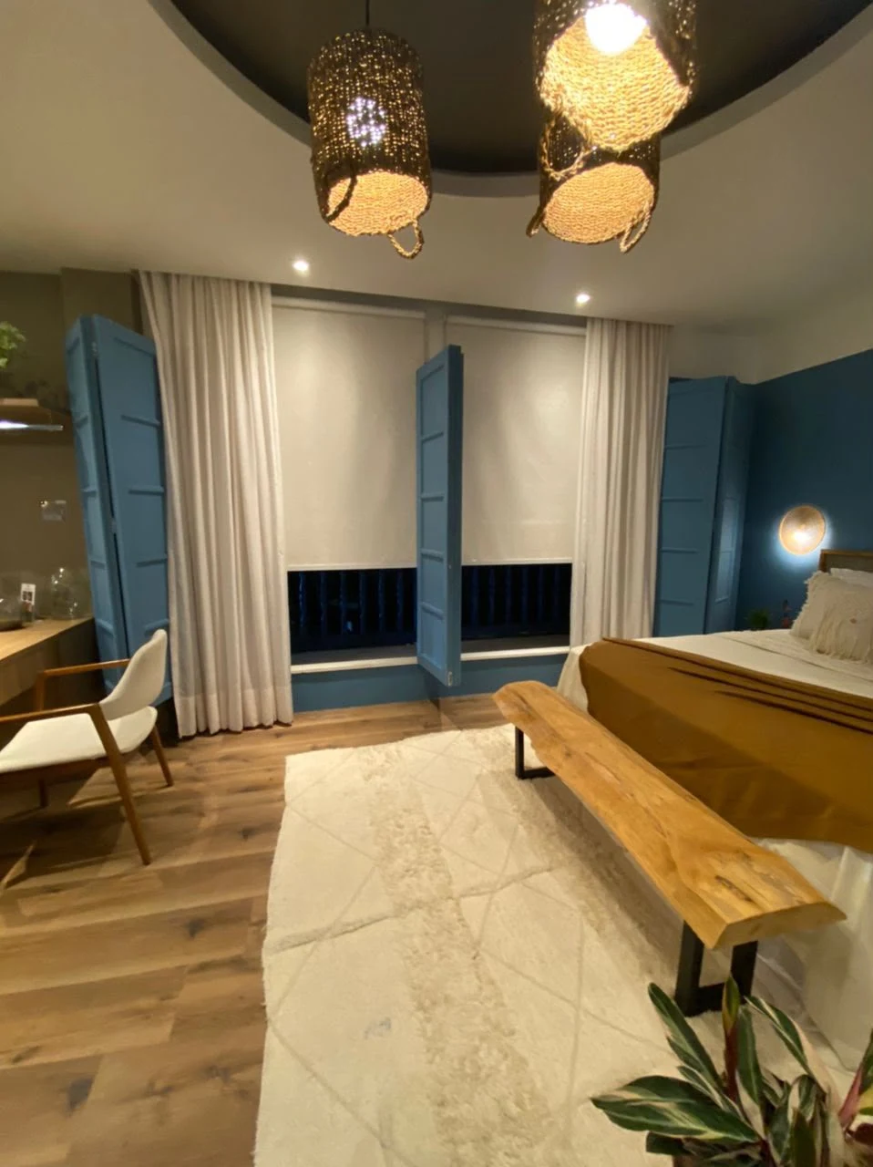 Quarto com janelas azuis, cortinas claras, persianas de rolo, tapete e banco de madeira aos pés da cama