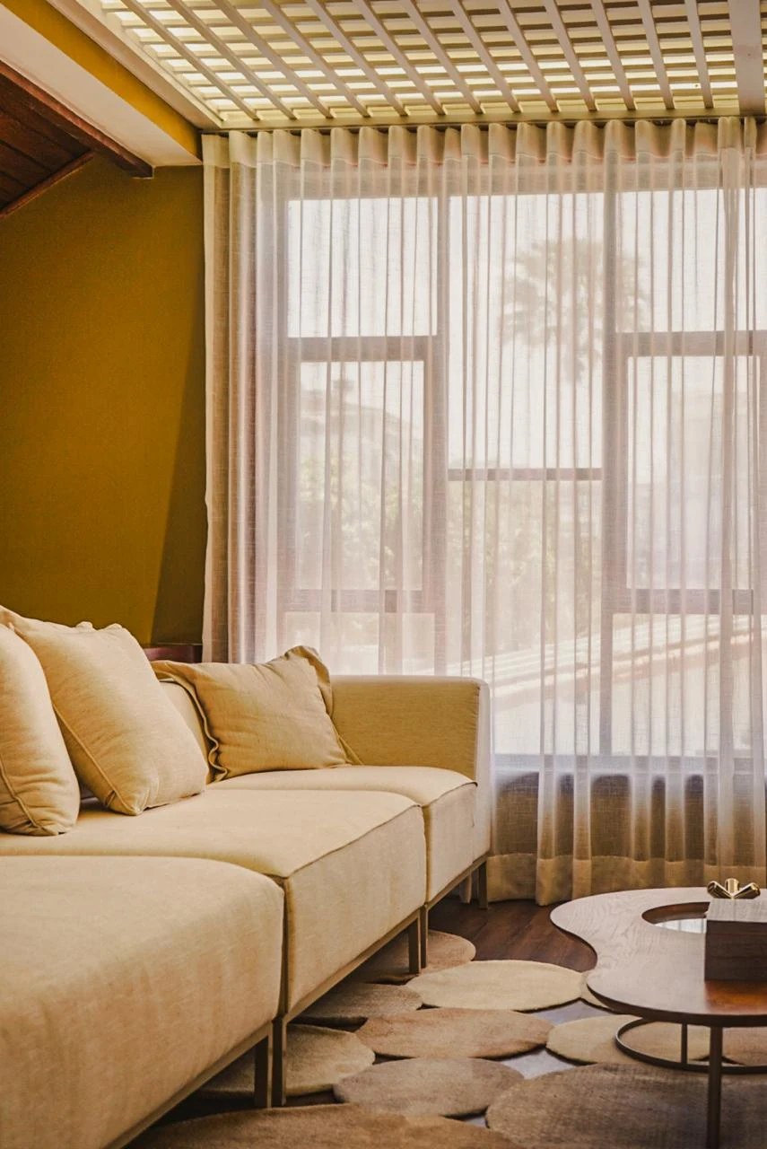 Sofá bege diante de cortinas leves do teto ao piso, com mesas de centro e tapete orgânico